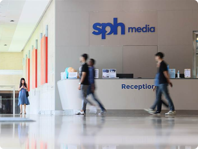 The image shows the reception area of SPH Media, a media company. The reception desk is positioned against a wall with the company's logo "sph media" prominently displayed above it in blue letters. Below the logo, the word "Reception" is written in smaller blue letters. Several people are walking past the reception area, and one person is standing near the desk. The floor is reflective, and there are colorful panels on the left wall of the hallway leading to this area.