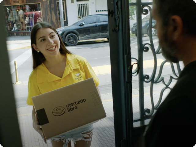 A smiling delivery worker wearing a yellow shirt hands a Mercado Libre package to a person standing at the door.